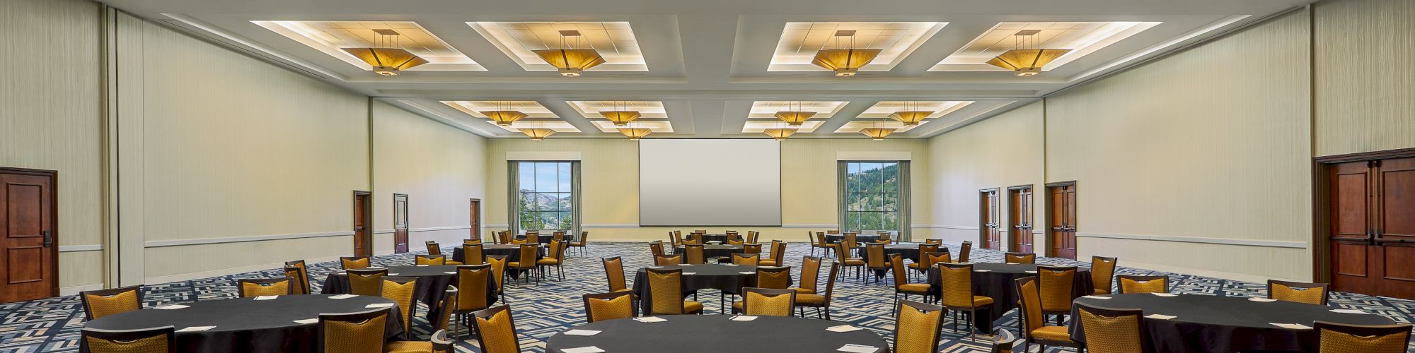A spacious, well-lit conference room with round tables and yellow cushioned chairs, patterned carpet, and modern ceiling lights.