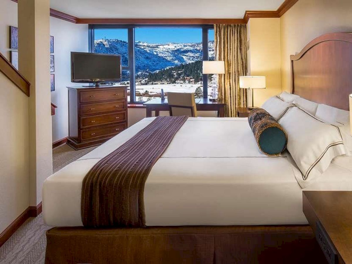 A cozy hotel room with a large bed, TV, desk, and a stunning view of snowy mountains through the window, creating a serene atmosphere.