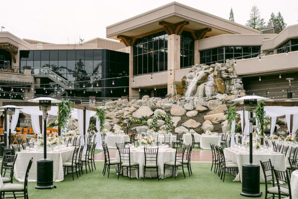 The image shows an elegant outdoor event setup with round tables, white tablecloths, black chairs, and decorative greenery, in front of a building and waterfall.