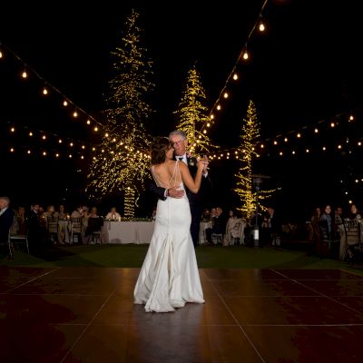 A couple is dancing together under string lights at an outdoor event, likely a wedding, with guests seated around them.