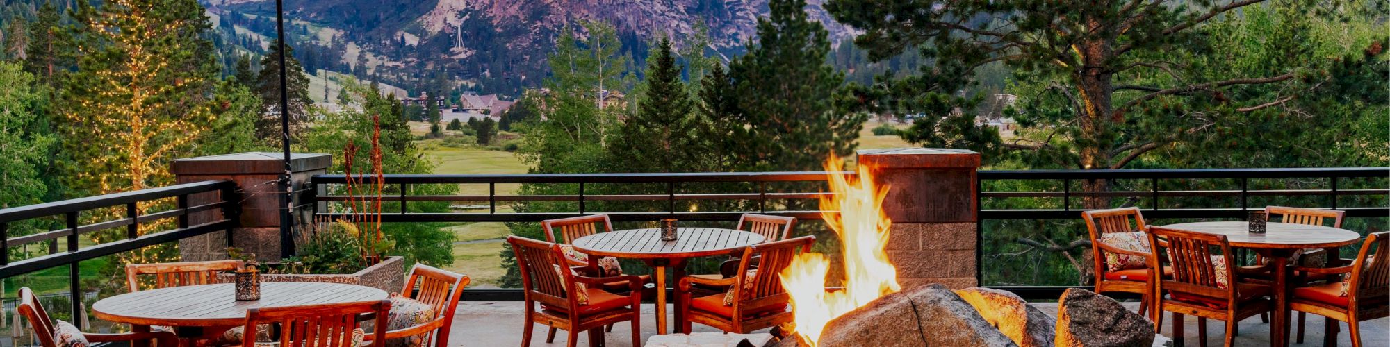 An outdoor patio with wooden tables and chairs, string lights, a stone fire pit, and a stunning mountain view in the background.