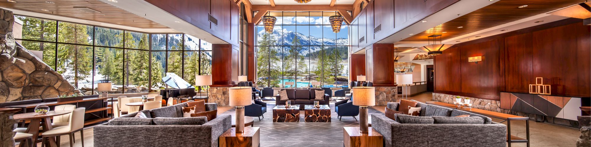 The image shows a spacious hotel lobby with modern furnishings, large windows offering scenic views, a high ceiling, and stylish pendant lights.