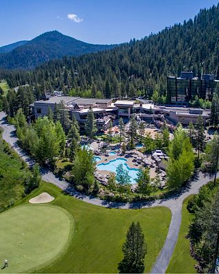 Aerial view of a resort nestled in lush green mountains, featuring a swimming pool, surrounding foliage, golf course, paths, and a serene lake.