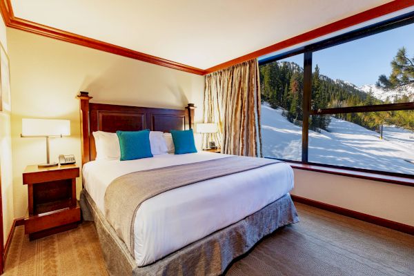 A cozy bedroom with a large bed, two bedside tables, lamps, and a window offering a snowy mountain view on a sunny day.