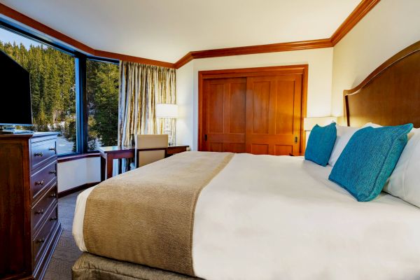 A cozy hotel room features a large bed with blue pillows, a TV, a dresser, and a window with a scenic view of trees and mountains.