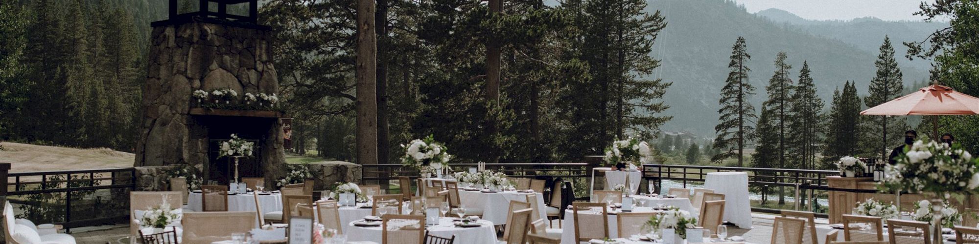 An outdoor event setup with round tables and chairs against a mountainous backdrop, decorated for a formal occasion with floral centerpieces.