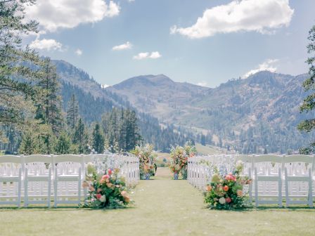 An outdoor wedding setup with white chairs and floral decorations, situated in a scenic mountain landscape under a partly cloudy sky.