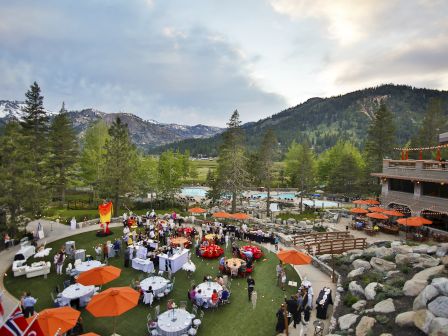 An outdoor event with tables, orange umbrellas, and various flags in a mountain setting, surrounded by trees and a building with a patio.