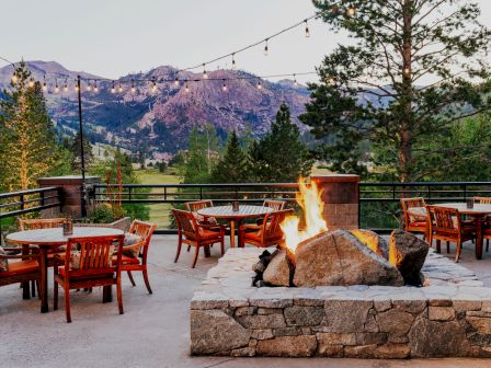 An outdoor seating area with wooden tables and chairs, a stone fire pit, string lights, surrounded by mountains and trees at sunset.