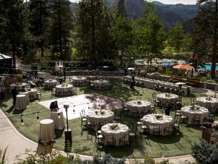 An outdoor event setup with round tables and chairs on a lawn, surrounded by trees and mountains in the background, under a partly cloudy sky.