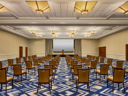 A conference room with rows of chairs facing a podium and screen, featuring modern lighting and geometric carpet design, ready for an event.