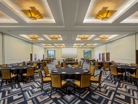The image shows a large, modern conference or banquet hall with round tables, yellow chairs, patterned carpeting, and ceiling lights.