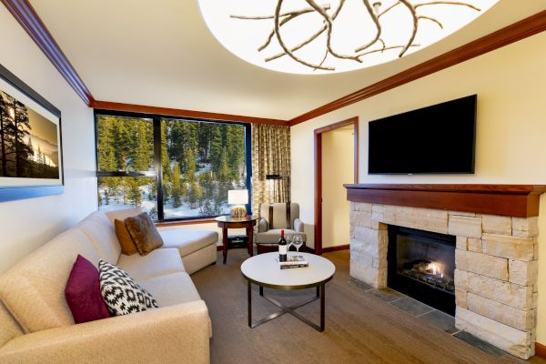 A cozy living room features a beige sofa, a fireplace, a wall-mounted TV, and a large window with a scenic view.