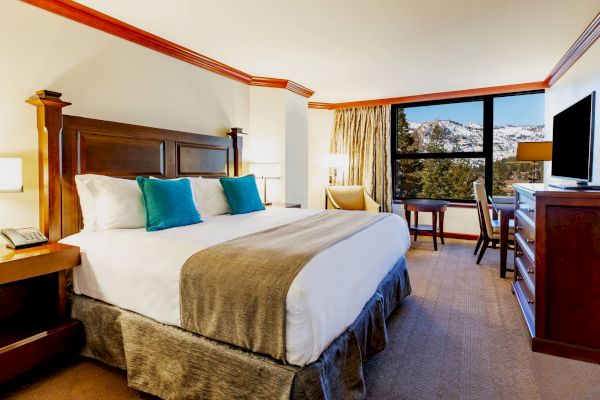 This image shows a hotel room with a large bed, wooden furniture, a television, and a window with a scenic view of a snowy mountain landscape.