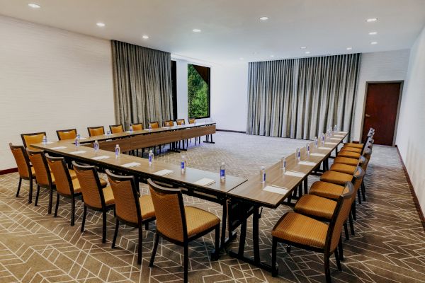 A conference room set up with U-shaped tables, chairs, notepads, and water bottles, with curtains and a door in the background ends the sentence.