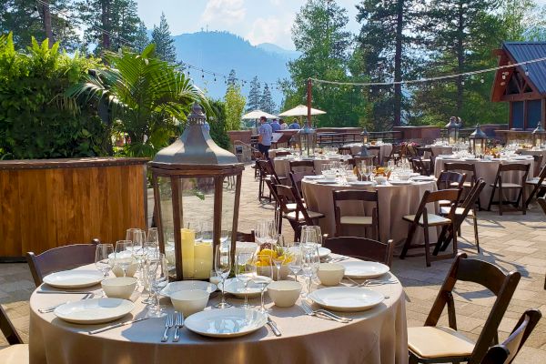 An elegantly set outdoor dining area with round tables, wooden chairs, a lantern centerpiece, and a mountain scenery in the background.
