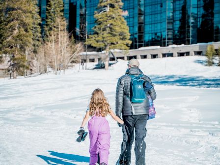 A person and a child in winter clothing walk hand in hand on a snowy path, with trees and a modern glass building in the background.