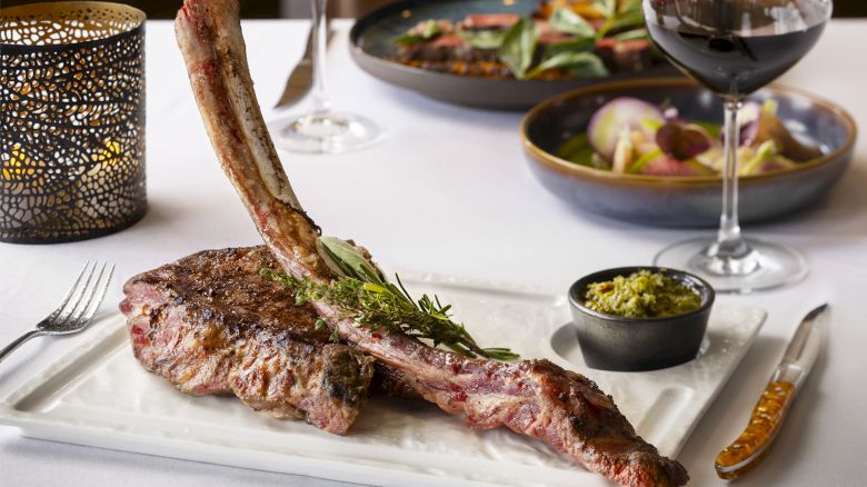 A plate with a large tomahawk steak garnished with herbs, a small bowl of sauce, a candle, wine glasses, and additional dishes in the background.