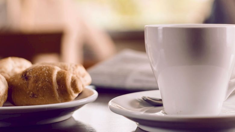 A white coffee cup on a saucer sits next to a plate of pastries on a table, with a rolled-up newspaper in the background.