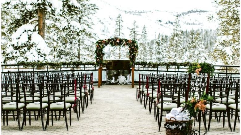 Snowy outdoor wedding setting with chairs, floral arch, and mountainous background, surrounded by snow-covered trees and serene winter scenery.