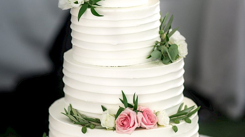 A three-tiered white wedding cake adorned with pink and white flowers and green leaves, placed on a table with plates and a gold cake server.