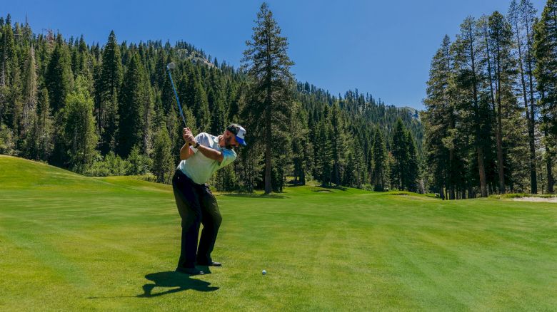 A golfer is mid-swing on a lush, green golf course surrounded by tall trees and mountains under a clear blue sky.