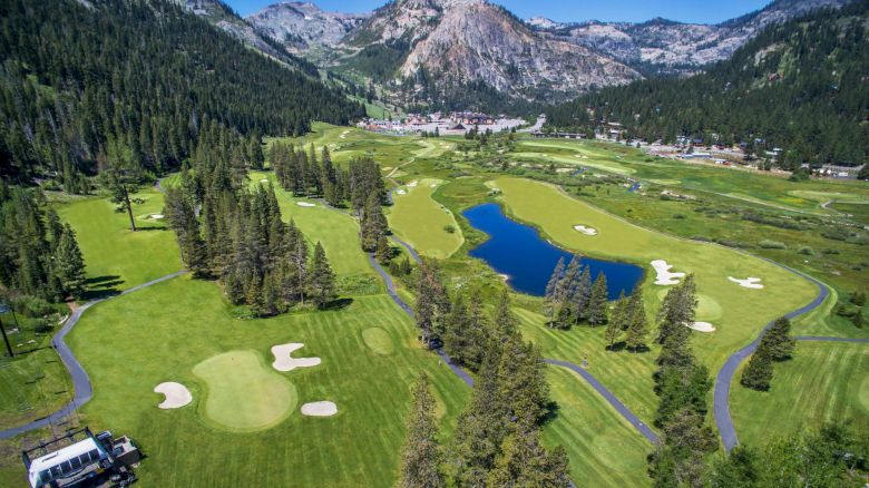 A picturesque golf course situated in a mountainous landscape, featuring greens, sand bunkers, a winding path, a pond, and lush trees.