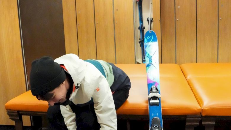 A person is sitting on an orange bench, adjusting their ski boot with a colorful ski leaning against the bench in a wooden room.