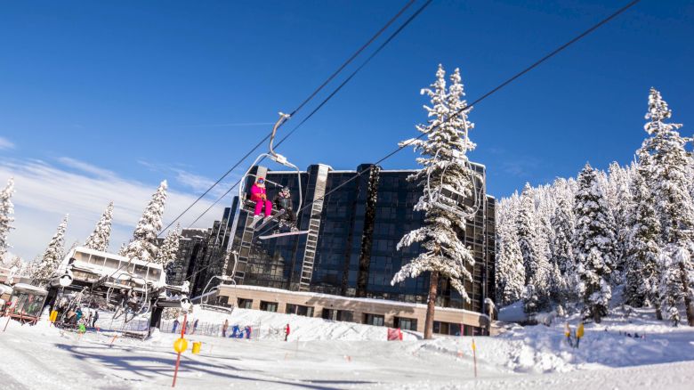 The image shows a snowy ski resort with people on a ski lift, a large building, and snow-covered trees under a clear blue sky.