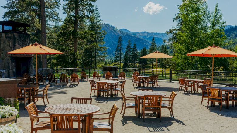 An outdoor seating area with wooden tables, chairs, and orange umbrellas is set against a scenic mountain view with tall trees and a clear sky.