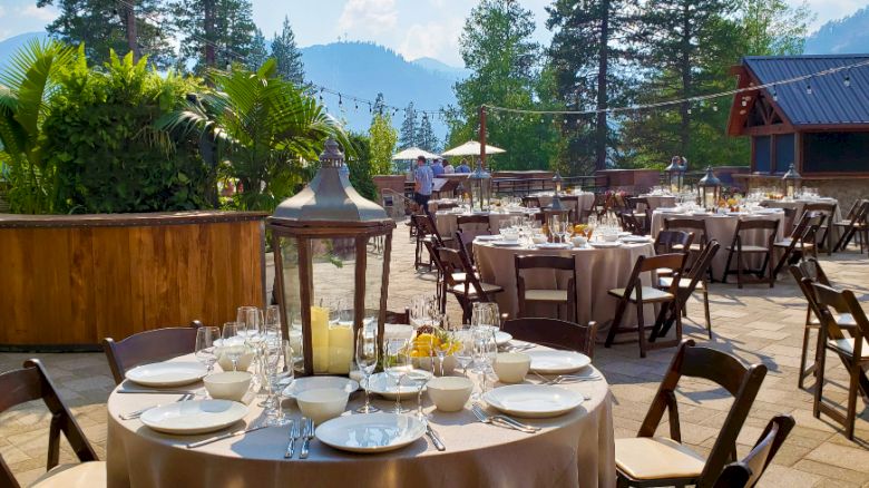 An outdoor event setup with round tables, chairs, and table settings, surrounded by trees and mountains in the background.