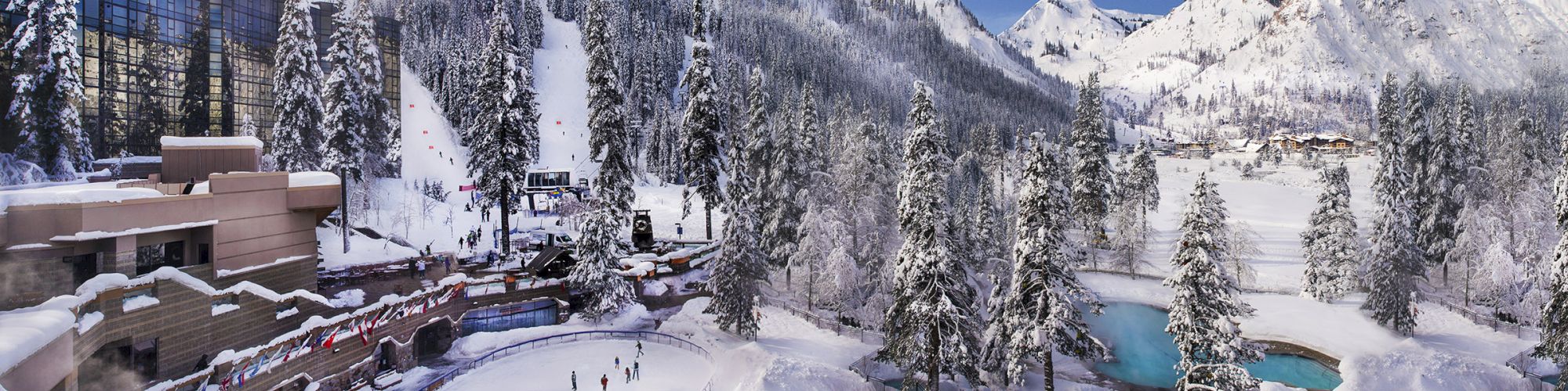 A snowy mountain resort with ski slopes, tall trees, buildings, and a blue sky with scattered clouds in the background, surrounded by snow-covered peaks.