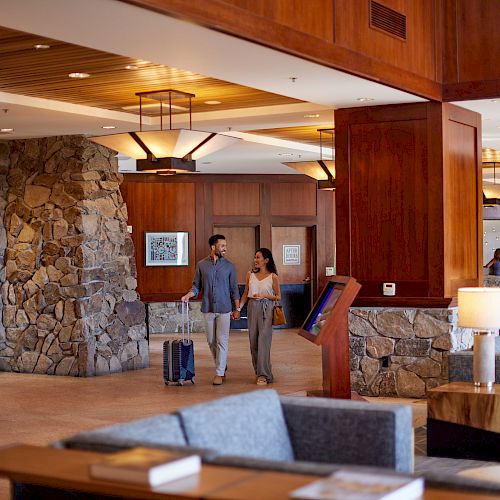 A couple walks through a hotel's stylish, cozy lobby with modern decor, stone accents, and wooden paneling, pulling luggage near the front desk.