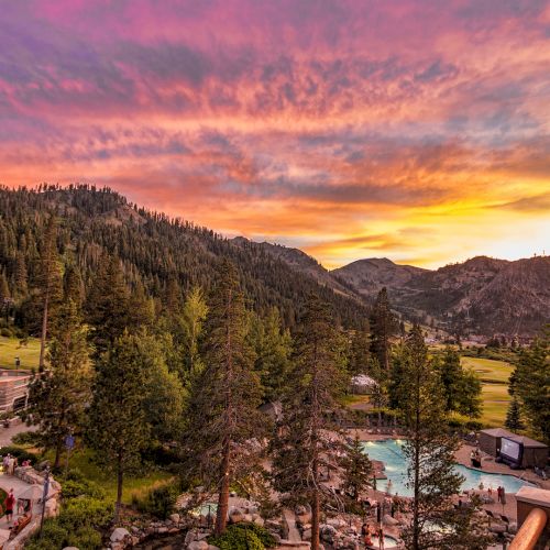 A scenic view of a resort at sunset with a colorful sky, surrounded by trees and mountains. The image also includes a swimming pool and buildings.