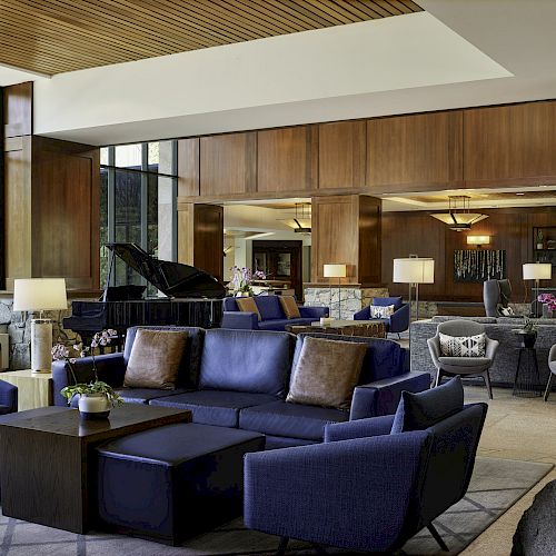 A spacious, contemporary living area features wooden accents, a grand piano, modern blue seating, large windows, and ambient lighting.