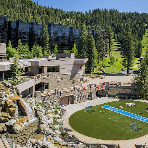 The image displays a resort complex nestled in a lush forested area, featuring a large lawn, cascading waterfall, and modern buildings.