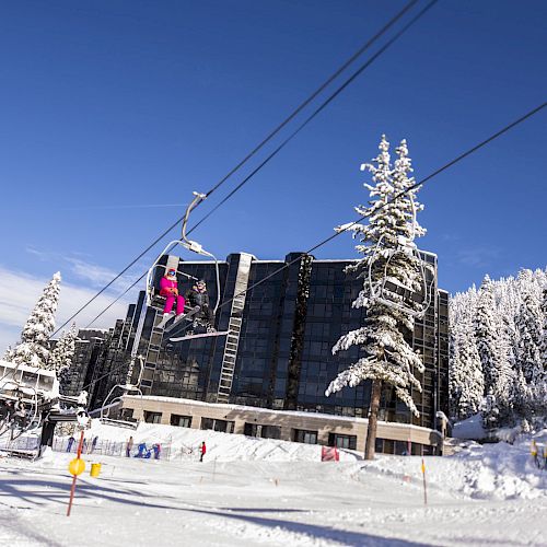 A snowy mountain resort with people on ski lifts, tall snow-covered trees, and a large building in the background under a clear blue sky.