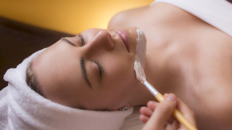 A person with a towel wrapped around their head, lying down and receiving a facial treatment with a brush applying a cream to their face.