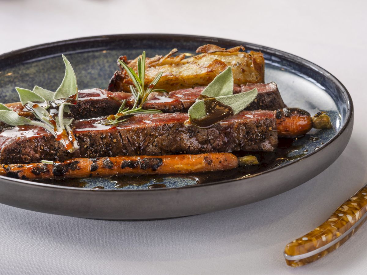A plate of sliced steak garnished with herbs, accompanied by roasted carrots and potatoes, placed on a dark round plate.