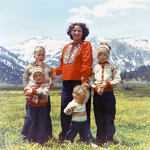 A woman and four children, all dressed in matching outfits, stand in a meadow with snow-capped mountains in the background.
