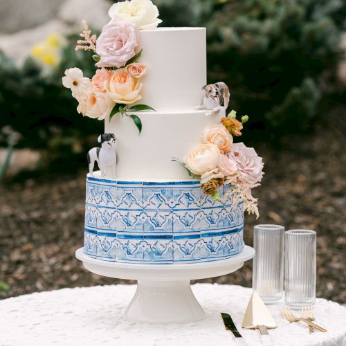 A three-tiered white wedding cake with blue patterns and floral decorations, accompanied by two champagne flutes on a white lace-covered table.
