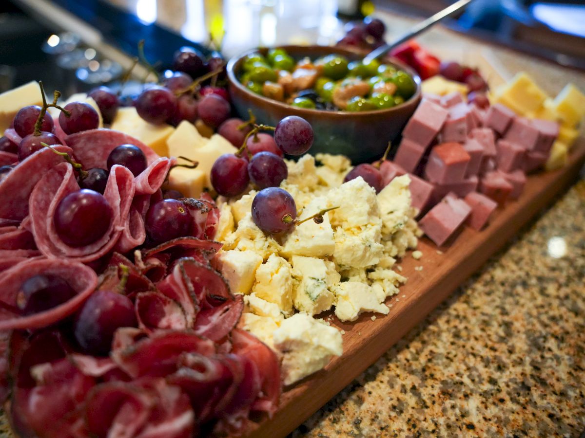 A charcuterie board featuring grapes, various cheeses, cubed ham, salami, and a bowl of mixed olives and nuts, all on a granite counter.
