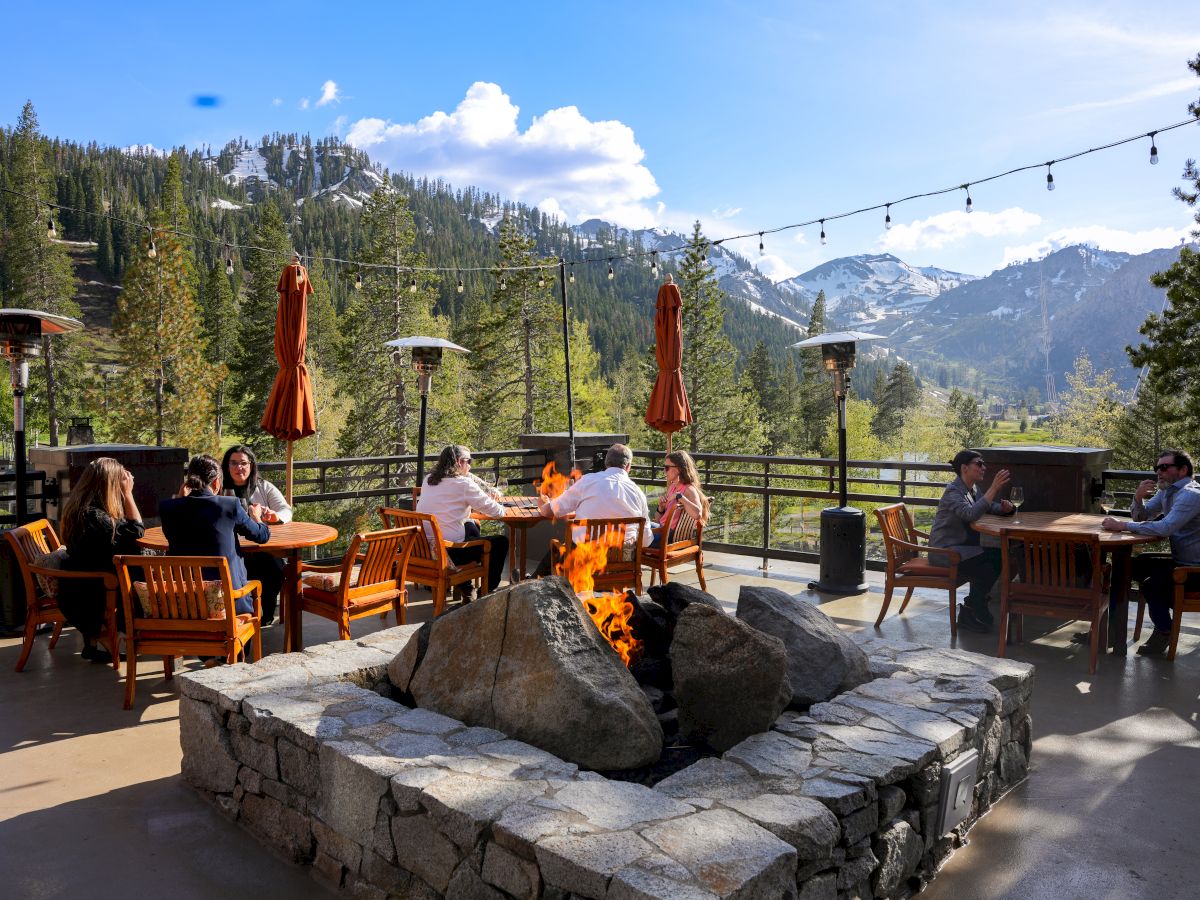 People sitting around tables and a fire pit on a mountain-view patio with string lights and umbrellas, surrounded by trees and snowy peaks.