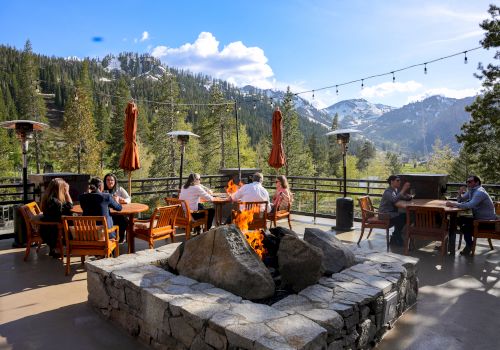 People sitting around a fire pit on an outdoor patio with mountains and trees in the background, under a clear sky with fairy lights strung above.