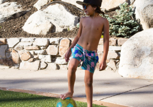 A child wearing shorts, sunglasses, and a hat stands on grass with one foot on a soccer ball, with rocks and plants in the background.