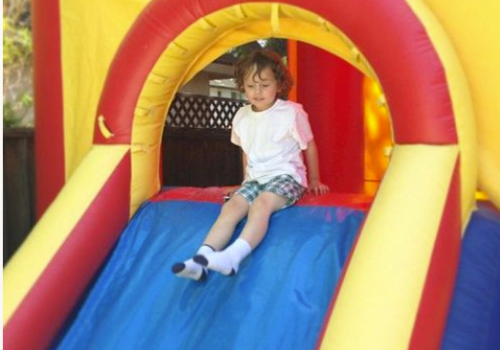 A child with curly hair, wearing a white shirt and checked shorts, is sliding down an inflatable slide in a colorful play area.