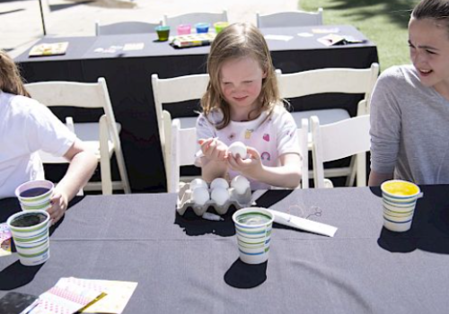 Children are sitting at a table with art supplies, eggs, and paint cups, appearing to be engaged in a craft activity outdoors.