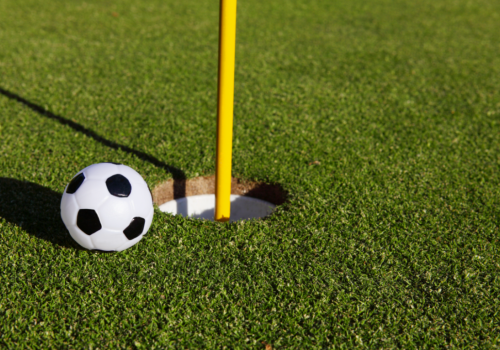 A small soccer ball is next to a hole on a golf course, with a yellow flagstick inserted in the hole. The scene is on a lush green putting green.